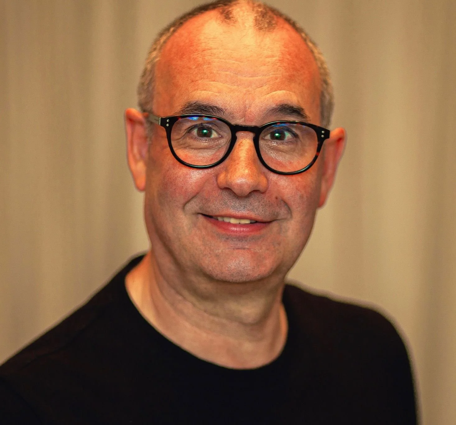 Close-up portrait of a smiling man with short hair, glasses, and a soft background.