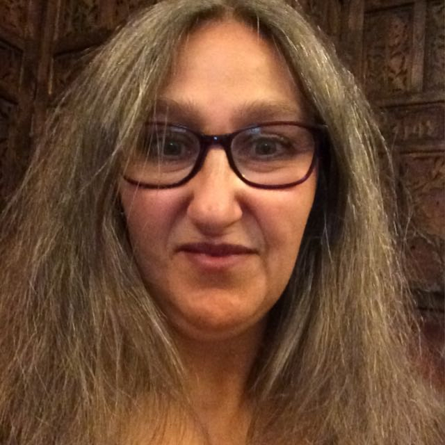 Close-up of a woman with long, wavy gray hair, wearing glasses, smiling slightly, with a wooden background.