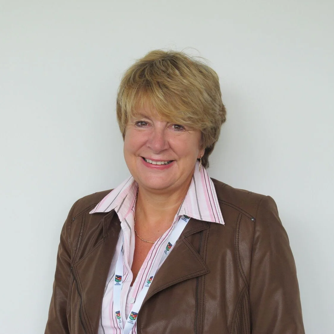 A smiling woman with blonde hair wearing a brown leather jacket and a pink striped shirt against a plain white wall.