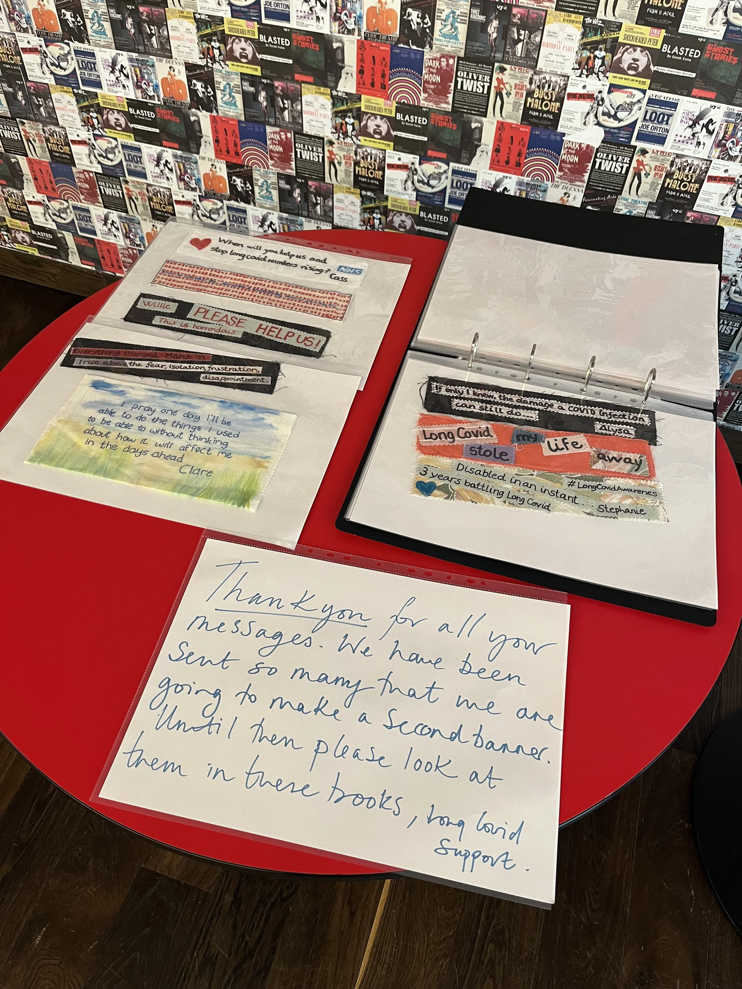 A red table with handwritten notes and letters, including messages of gratitude and support related to COVID-19, with a background wall covered in various book covers and posters.