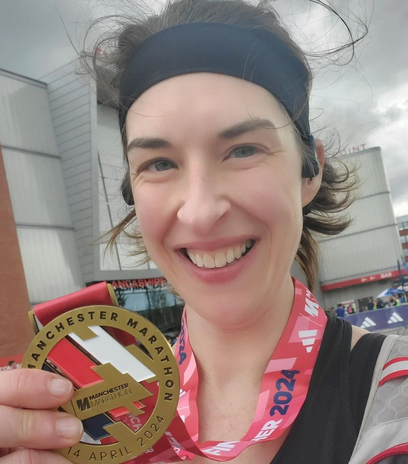 A woman smiling and holding a gold medal from the Manchester Marathon, wearing a black headband, with a cloudy sky and a building in the background.