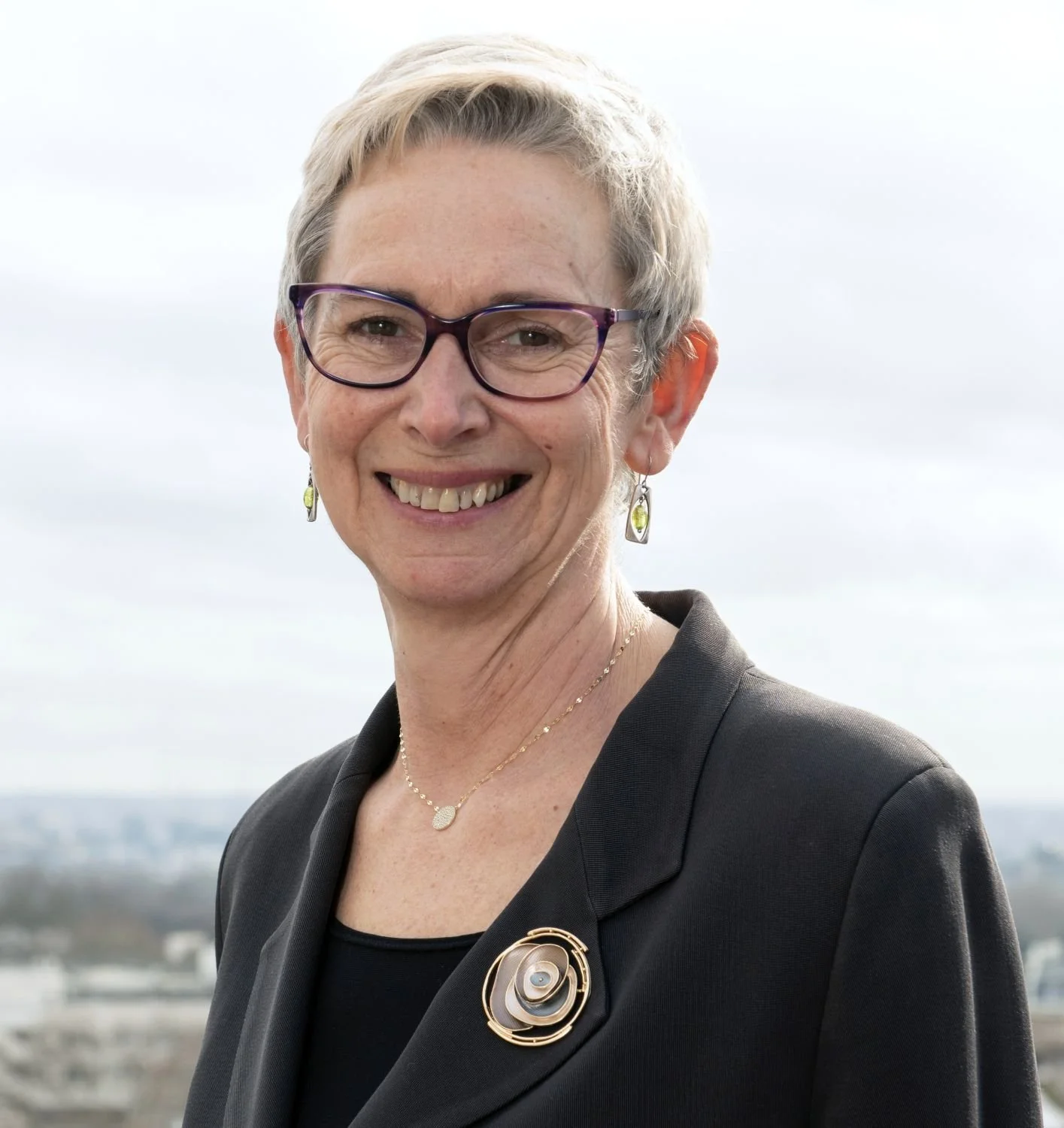 A smiling woman with short gray hair, glasses, earrings, and a necklace, wearing a black blazer with a decorative brooch, standing outdoors against a cloudy sky.