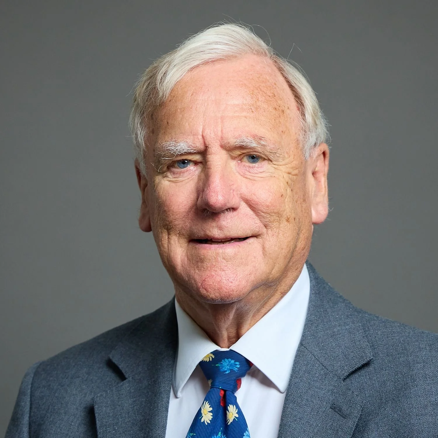 Portrait of an elderly man with white hair, blue eyes, wearing a gray suit, white shirt, and a tie with daisies, against a plain gray background.