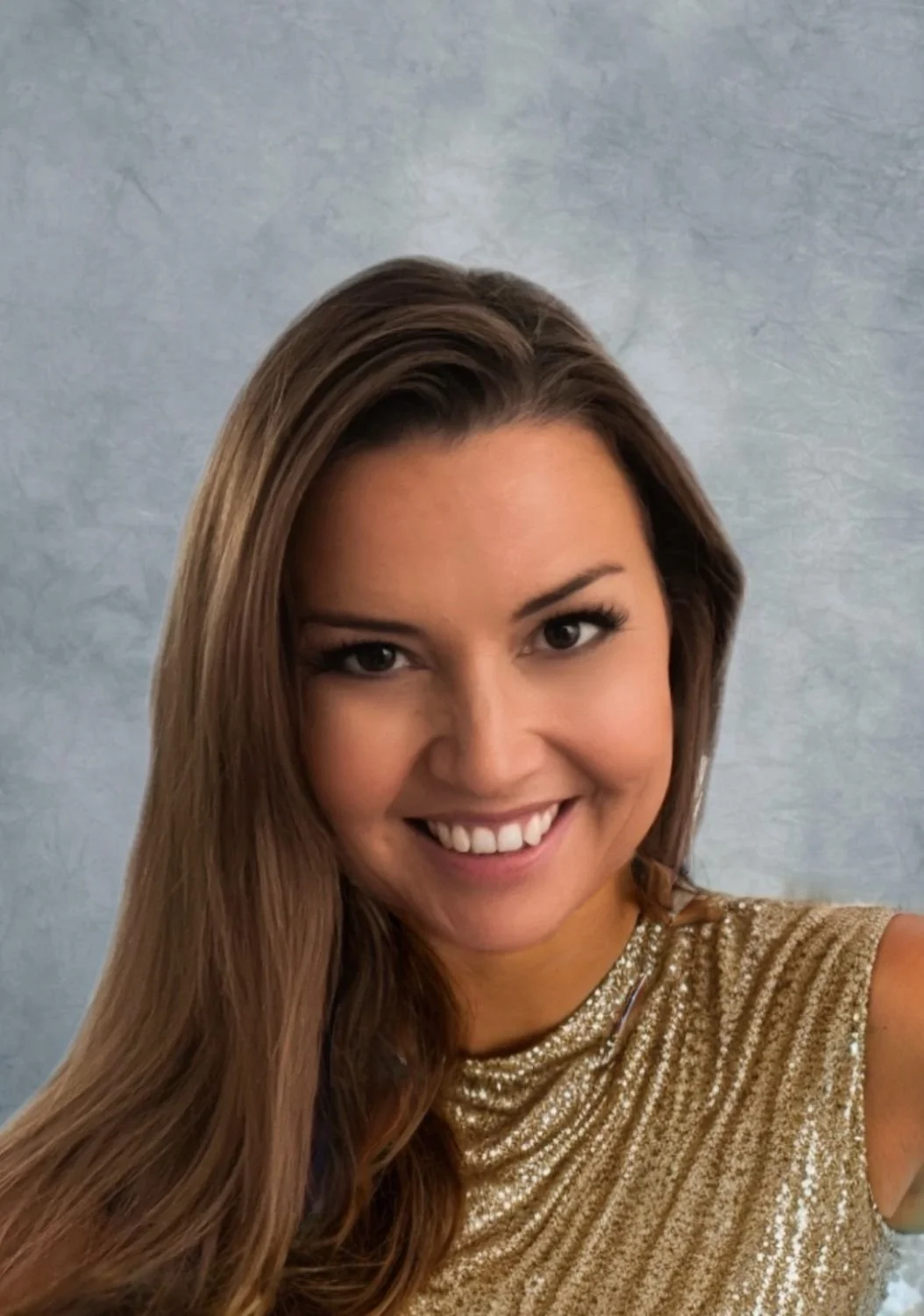 A smiling woman with long brown hair wearing a gold, shiny top, standing against a blue-gray background.