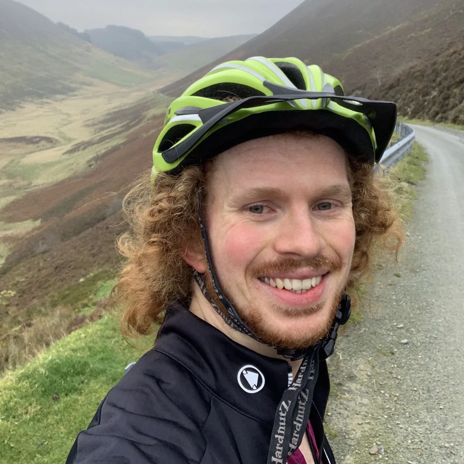 A smiling man with curly red hair and a beard wearing a black cycling jacket and a neon green helmet, taking a selfie on a mountain road with hilly terrain in the background.