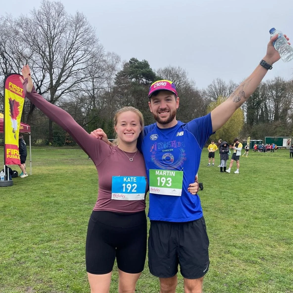 Two runners, a woman and a man, celebrate after a race at a park. They are smiling, with their arms raised. The woman wears a maroon long-sleeve shirt and black shorts, with bib number 192 labeled 'KATE.' The man wears a blue shirt, black shorts, and a pink cap, with bib number 193 'MARTIN.' In the background, there are other runners and event tents.