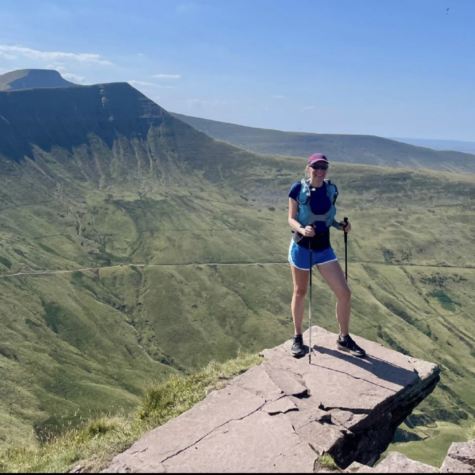 A woman hiking on a rocky formation in a lush green mountainous landscape, wearing a hat, sunglasses, shorts, and a backpack with hiking poles.