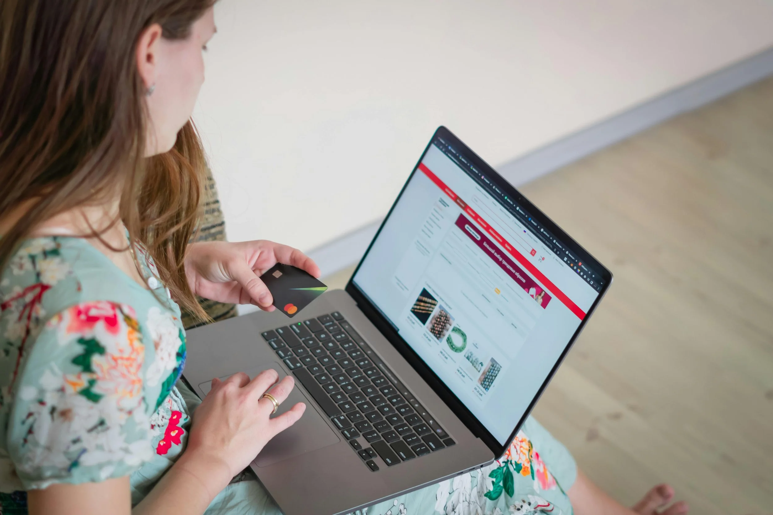 A woman with reddish hair wearing a multicolored floral dress is sitting on a beige couch, holding a black credit card in her right hand and using a laptop with a visible webpage open. The webpage appears to be a shopping or catalog site with images and red accents.