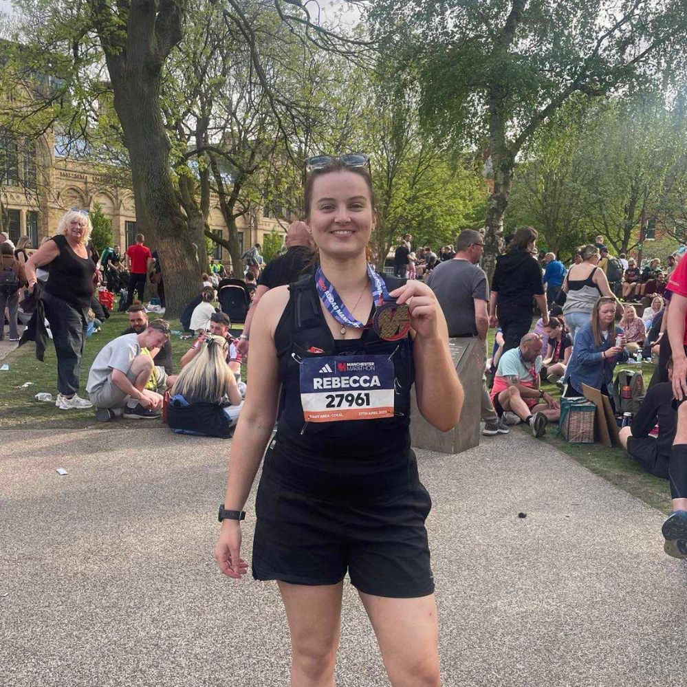 A young woman in black athletic wear, holding a medal and a race bib with the name Rebecca and number 27961, stands in a park after completing a marathon with people gathered around.