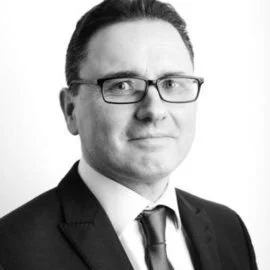 A professional headshot of a man with glasses, wearing a suit and tie, against a plain background.