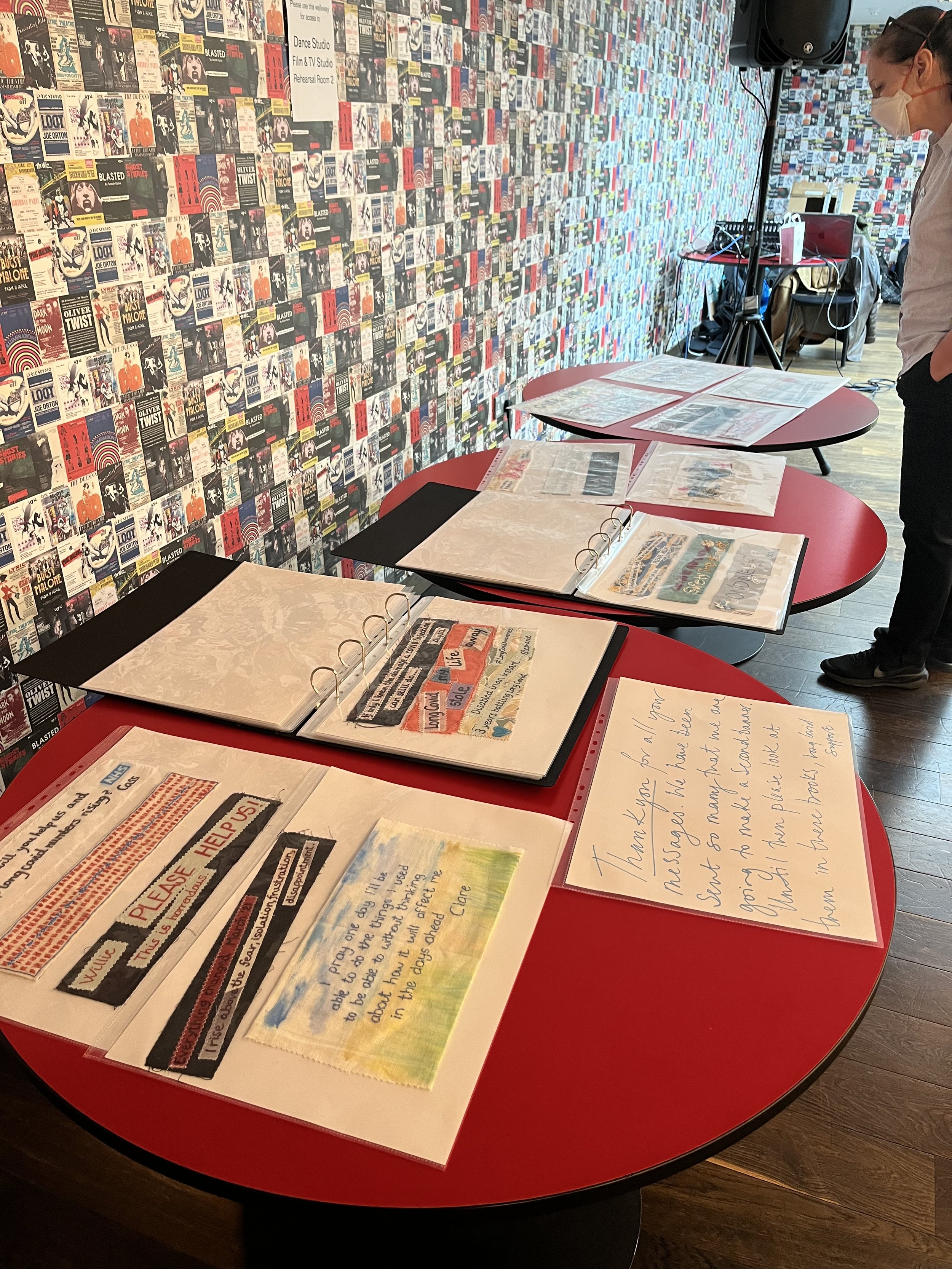A red circular table with open binders and handwritten notes inside, set in front of a wall covered with numerous posters. A person wearing glasses and a mask looks at the display, with audio equipment and a speaker nearby.