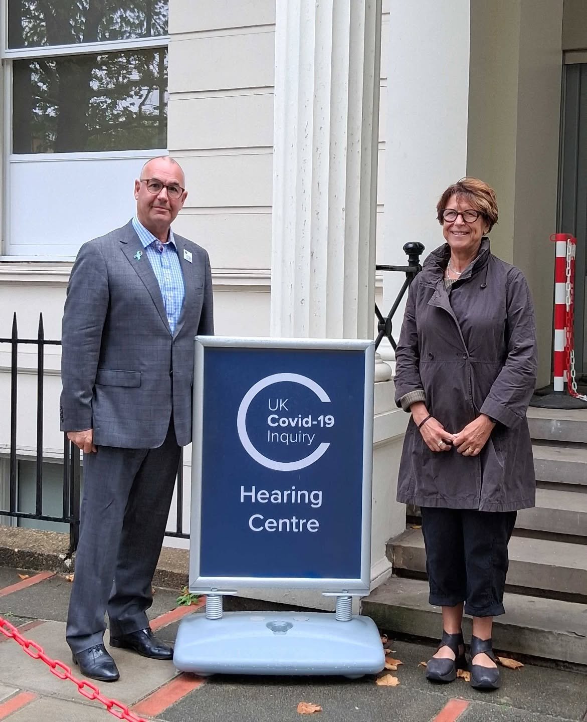 A man and a woman standing outside a building next to a sign that reads 'UK Covid-19 Inquiry Hearing Centre.' The man is wearing a gray plaid suit and glasses, and the woman is wearing a dark coat and glasses. They are both smiling and facing the camera.