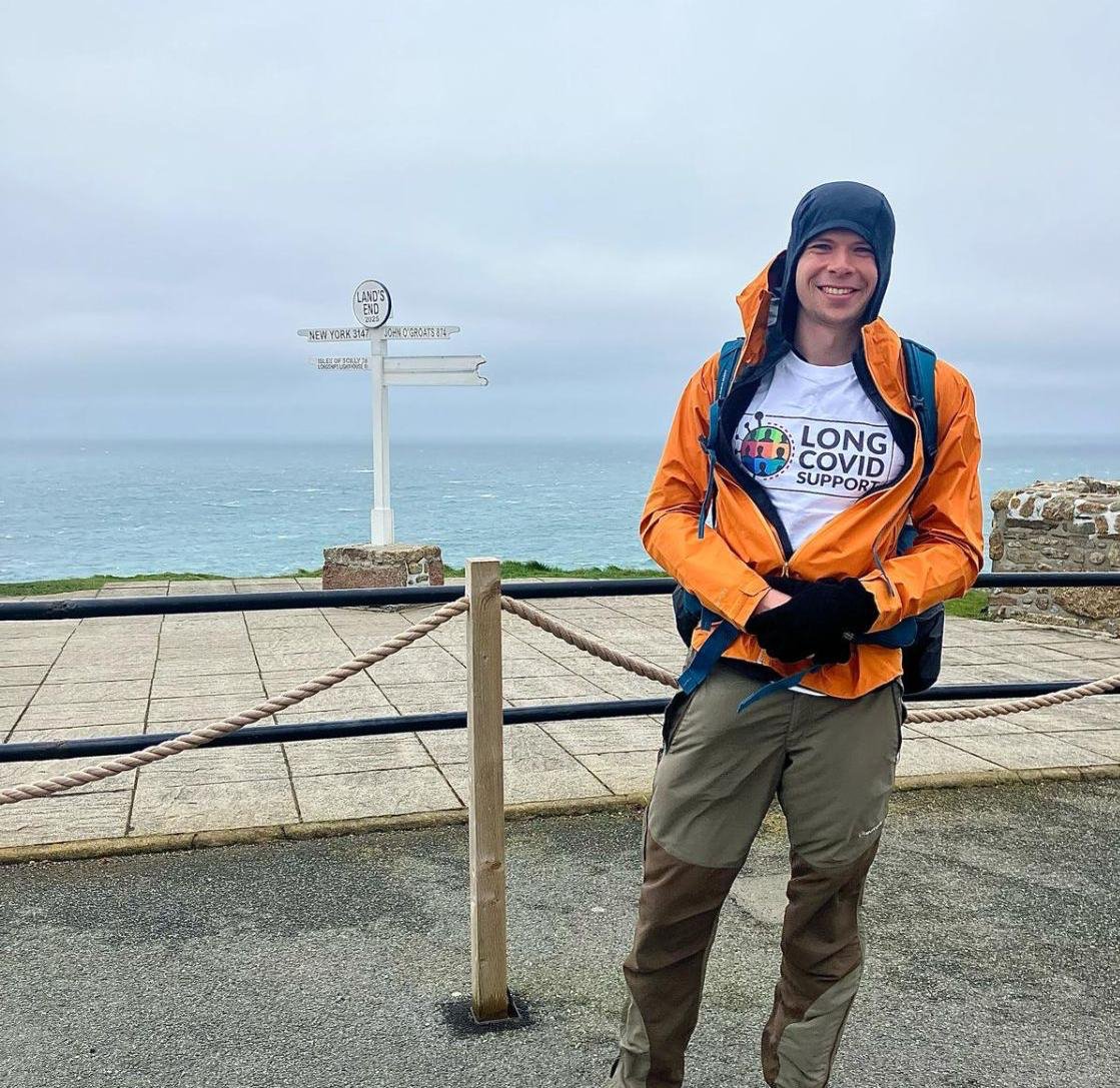 Man smiling at a seaside lookout point, wearing an orange rain jacket, gray pants, and a white t-shirt supporting Long COVID awareness, with a signpost in the background indicating the edge of land and the Atlantic Ocean.