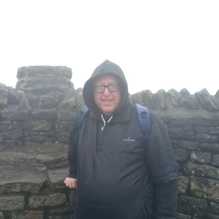 Man with glasses wearing a black waterproof jacket and hood, smiling, standing in front of a stone wall with foggy weather.