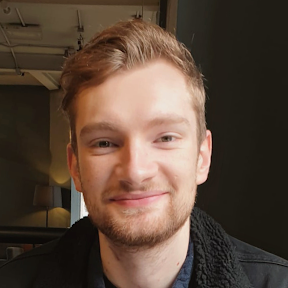 A young man with light brown hair and a slight beard, smiling softly at the camera