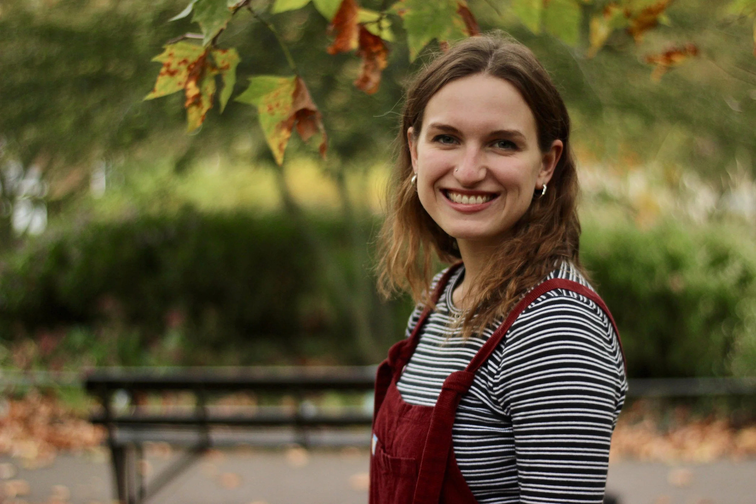 A young woman with brown hair, earrings, and a nose piercing, smiling while standing outdoors near a bench with autumn leaves in the background.