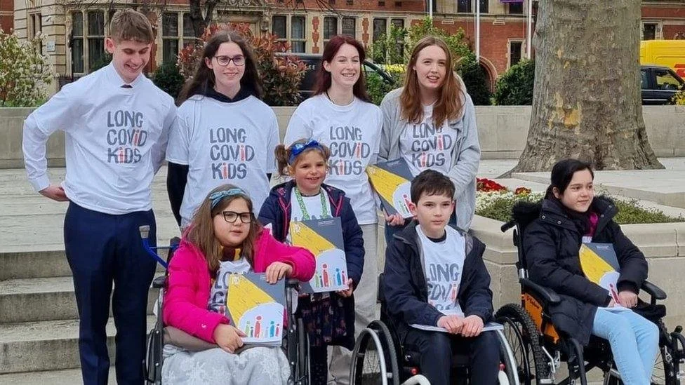 Group of children, some in wheelchairs, and teenagers standing behind them, wearing shirts that say "Long COVID Kids," posing outdoors in front of trees and buildings.
