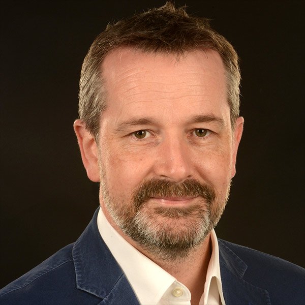 A middle-aged man with short brown hair, a beard, and facial hair, wearing a navy blazer and a white collared shirt, against a dark background.