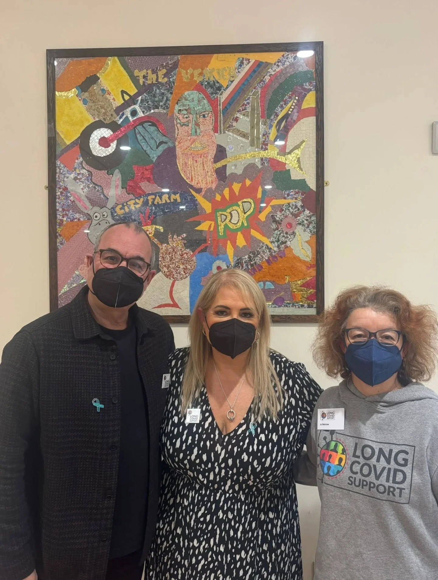 Four people standing in front of a colorful mosaic mural, all wearing face masks. The person on the right is wearing a gray shirt with "Long COVID Support" logo, the woman in the middle has a black and white patterned dress, the man on the left has glasses and a black suit, and the woman on the right has curly hair and glasses.