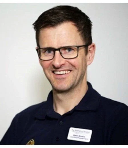 A man wearing glasses and a navy blue polo shirt with a name tag, smiling at the camera.