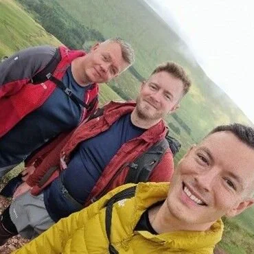 Three men hiking outdoors with green hills in the background, smiling at the camera.