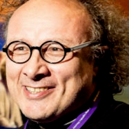 A man with curly hair, glasses, and a broad smile, wearing a black jacket.