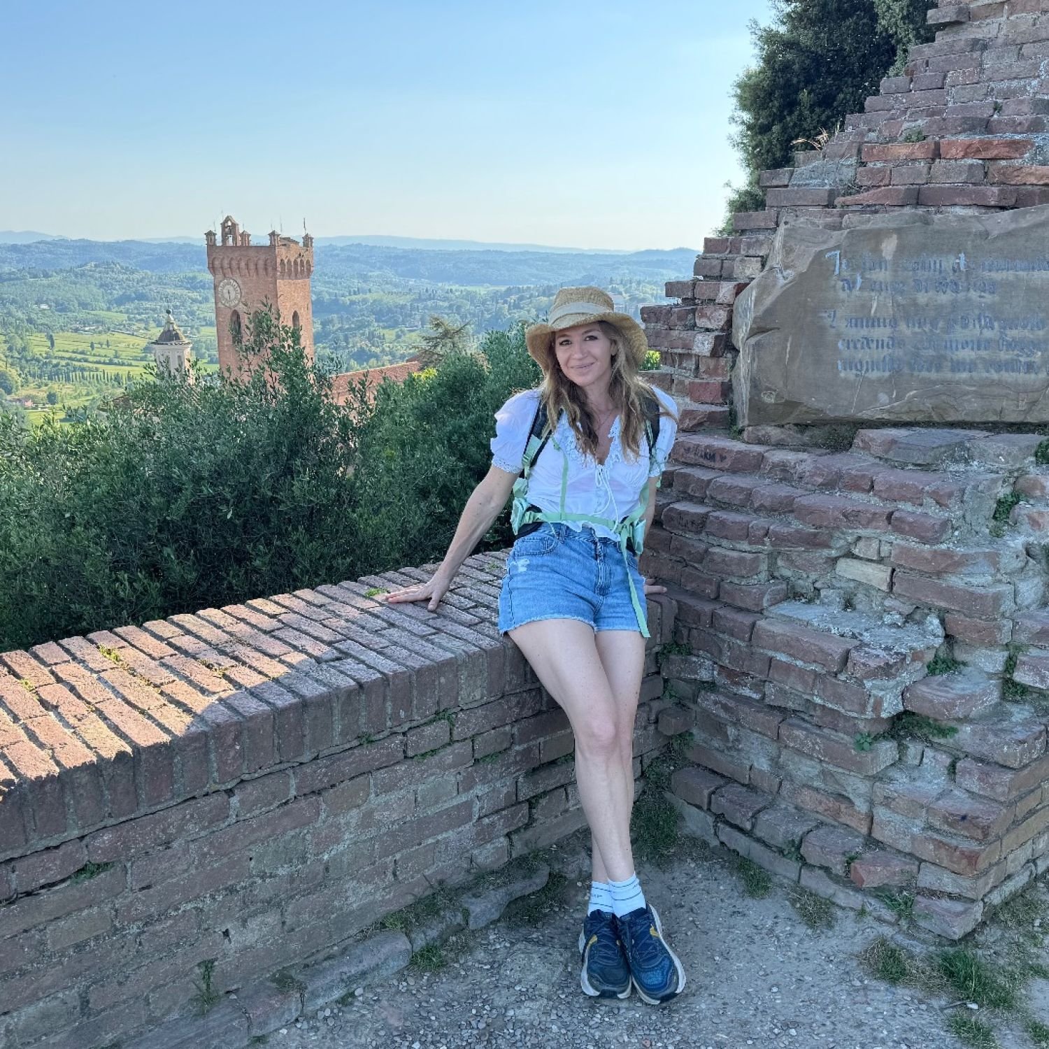A woman in casual summer clothing with a hat sitting on a brick wall in front of a scenic landscape with a tower, green hills, and trees
