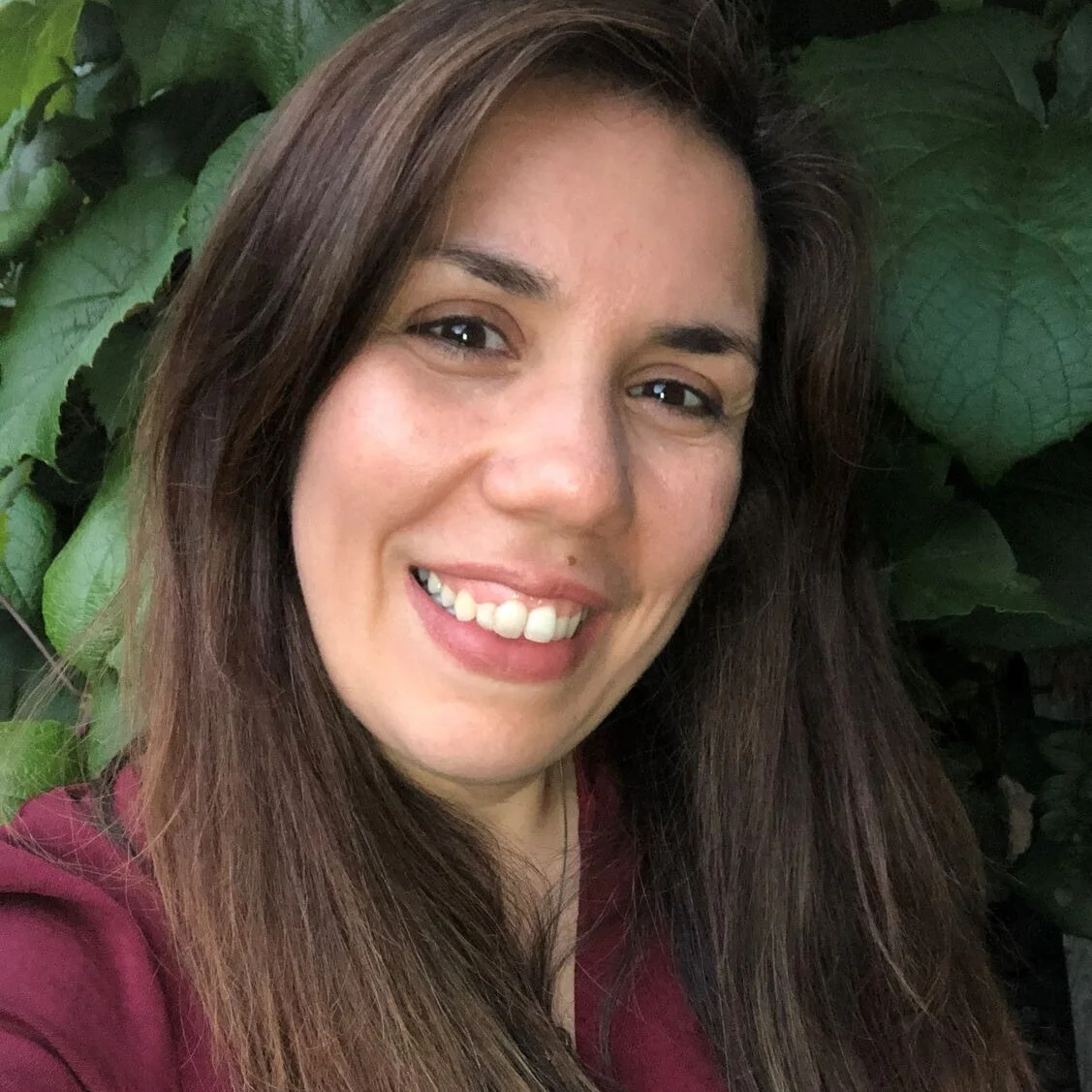 A smiling woman with long brown hair taking a selfie in front of green leaves.