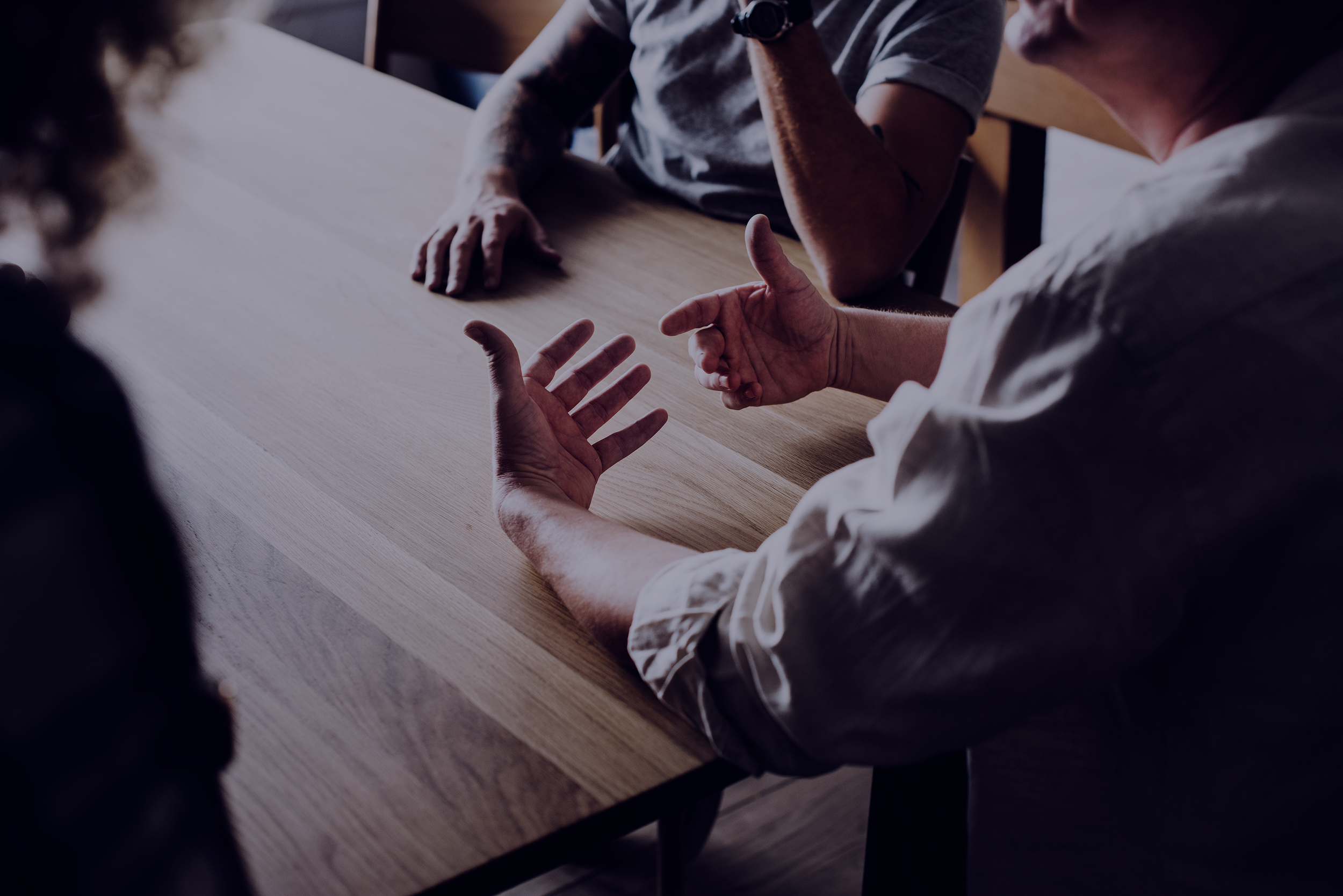 People engaged in a discussion at a wooden table, with focus on their hands and arms.