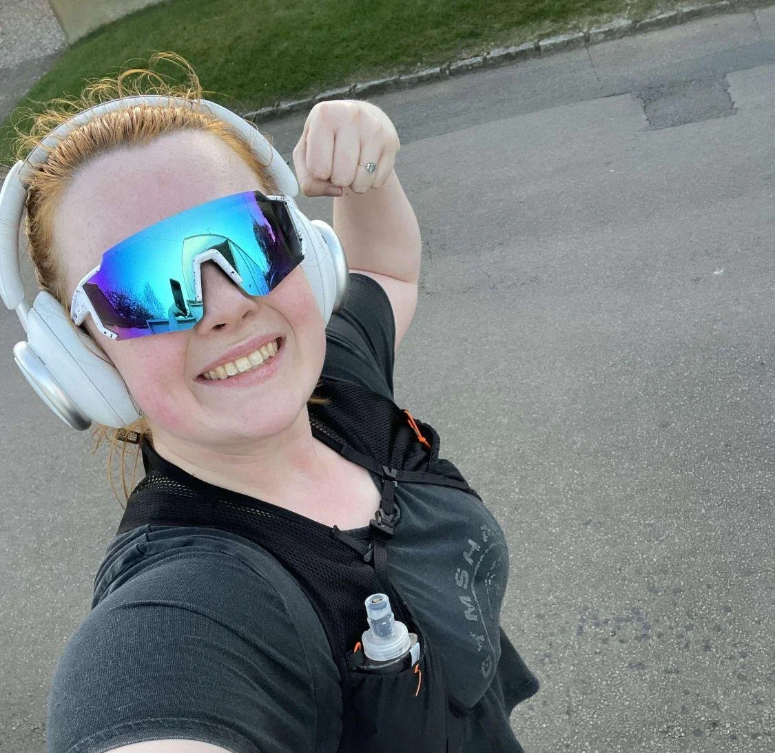 A woman with red hair wearing reflective sport sunglasses, large white headphones, a black athletic shirt, and a hydration pack, smiling while taking a selfie outdoors on a paved road.