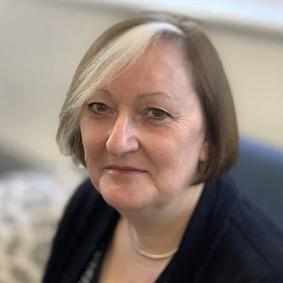 Close-up of a woman with short gray hair, wearing a dark blazer and a necklace, in an indoor setting.