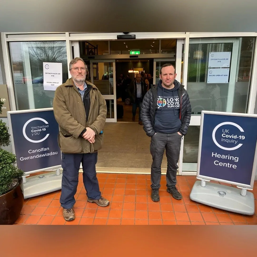 Two men standing outside a healthcare building with signs indicating a UK Covid-19 Inquiry and Hearing Centre; entrance to the building is behind them with people walking inside.