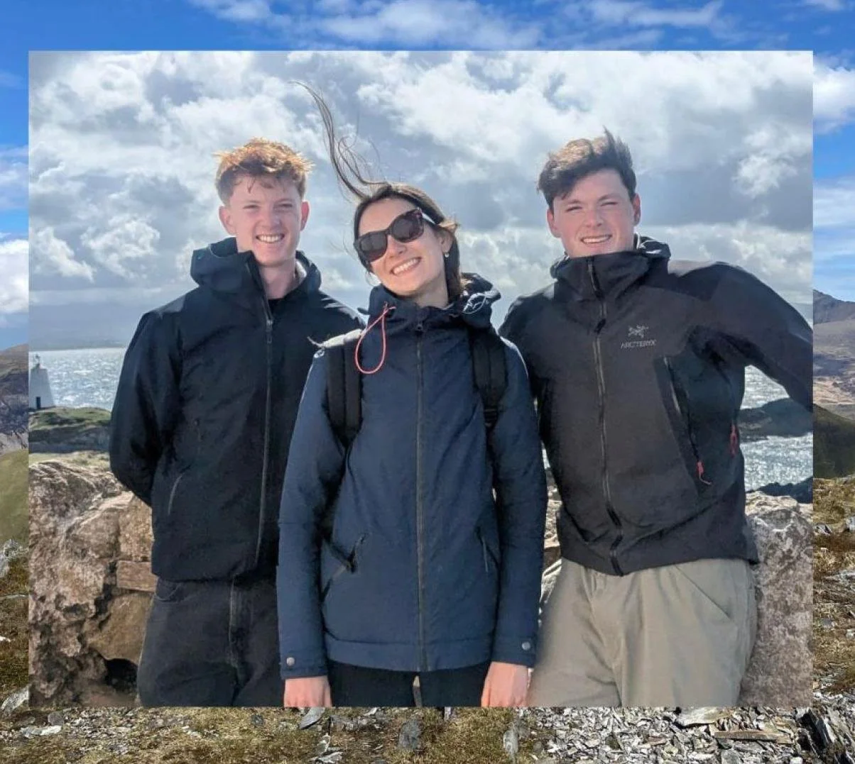 Three people standing outdoors on rocky terrain, smiling, wearing outdoor jackets, with a cloudy sky and water in the background.