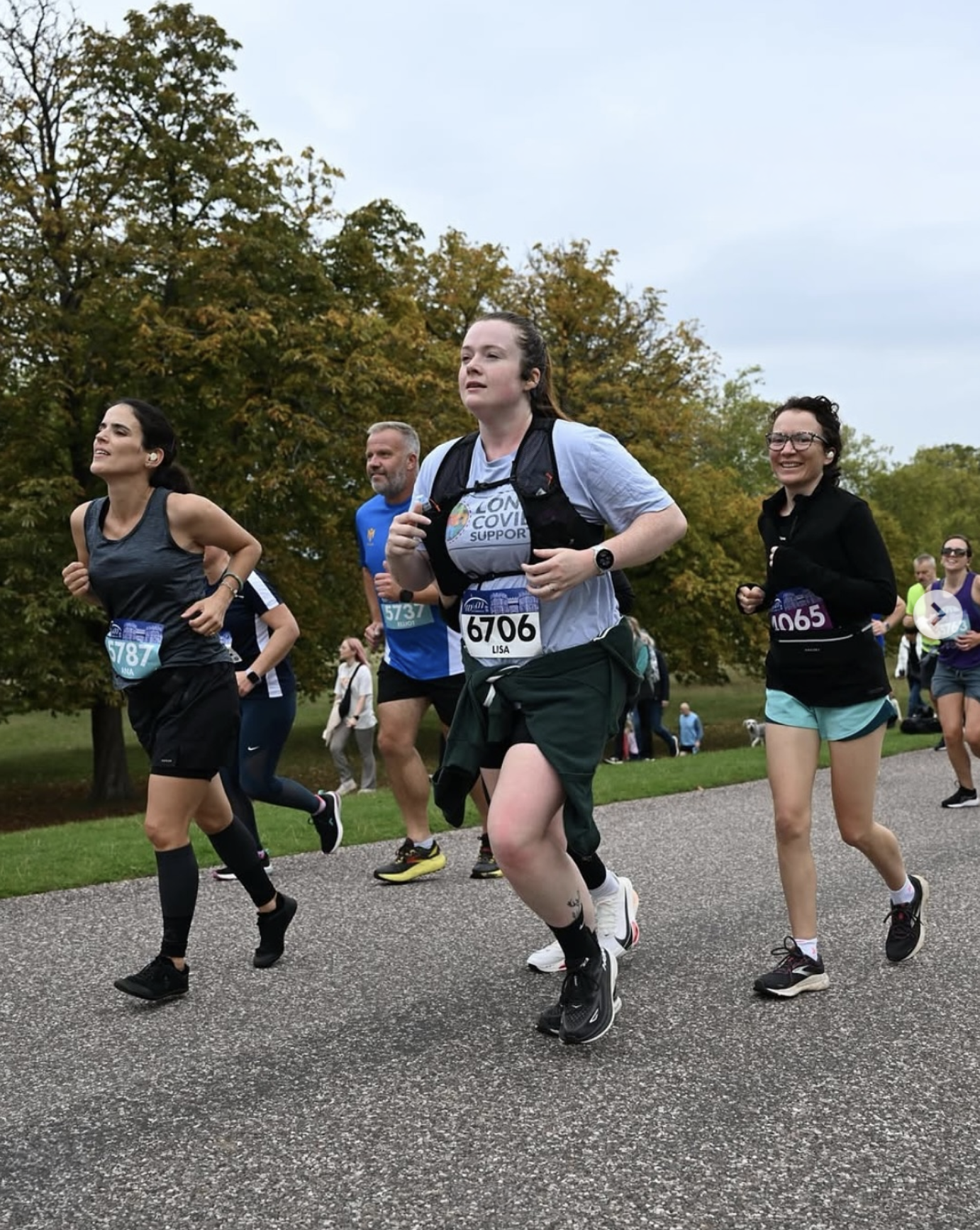 People running in a park during a race or marathon, wearing athletic clothing and race bibs, with trees and an overcast sky in the background.