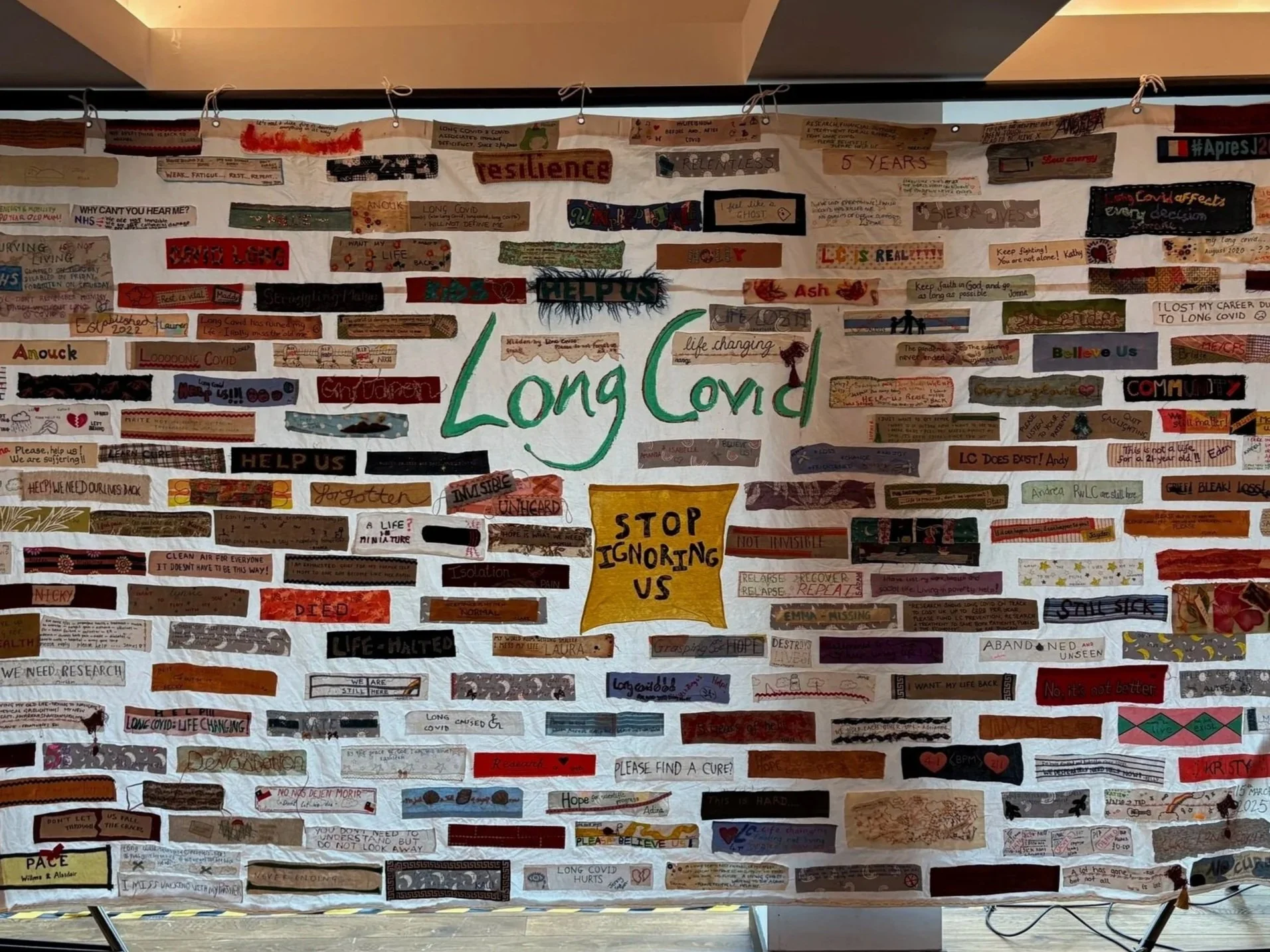 A large display board with numerous handwritten notes and messages related to long COVID, featuring the prominent words "Long Covid" in the center, and various words and phrases like "resilience," "help us," "stop ignoring us," and others in different colors and styles.