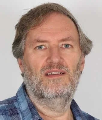 A middle-aged man with a beard and gray hair, wearing a blue plaid shirt, looking at the camera with a neutral expression.