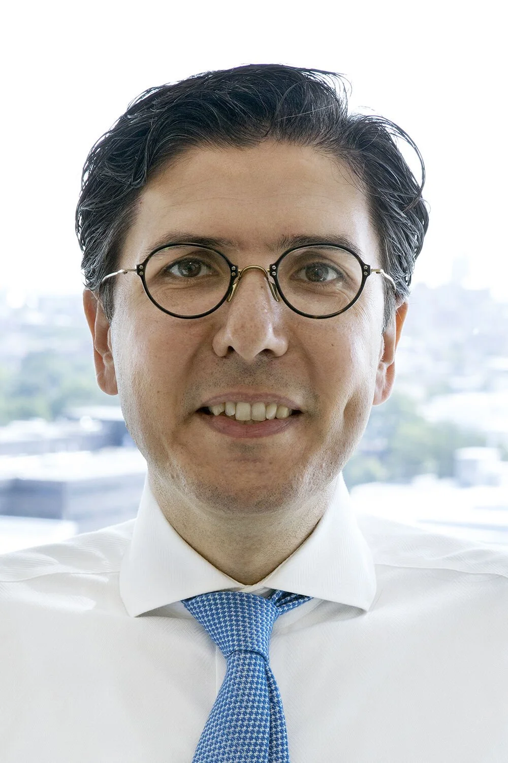Close-up of a man wearing glasses, white dress shirt, and blue tie, smiling slightly, with a cityscape background.
