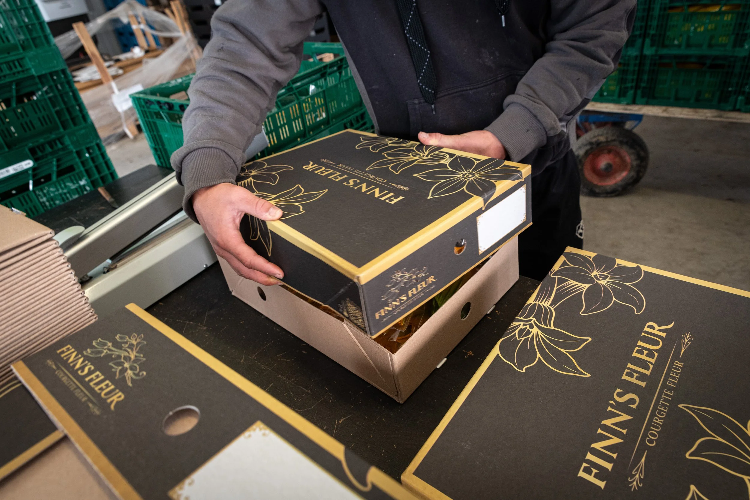 Person's hands holding a black box labeled 'Finn's FLEUR' with floral gold designs, surrounded by similar boxes and green plastic crates in a warehouse or storage area.