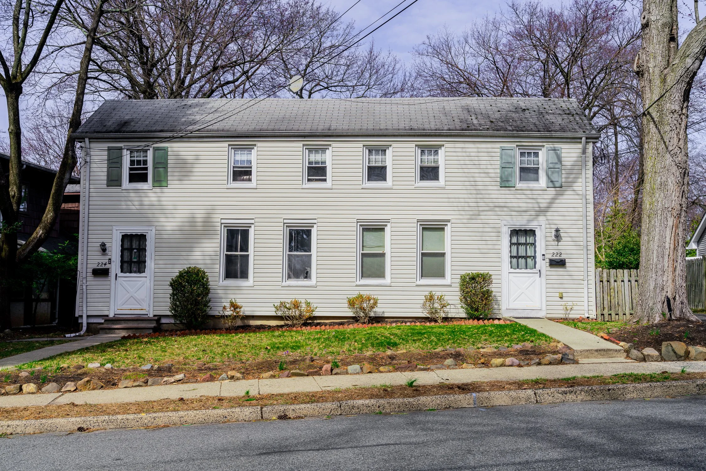 A white multi-story house with green shutters and a pitched roof. The front yard has small bushes, a concrete walkway, and a sidewalk, with large bare trees in the background.