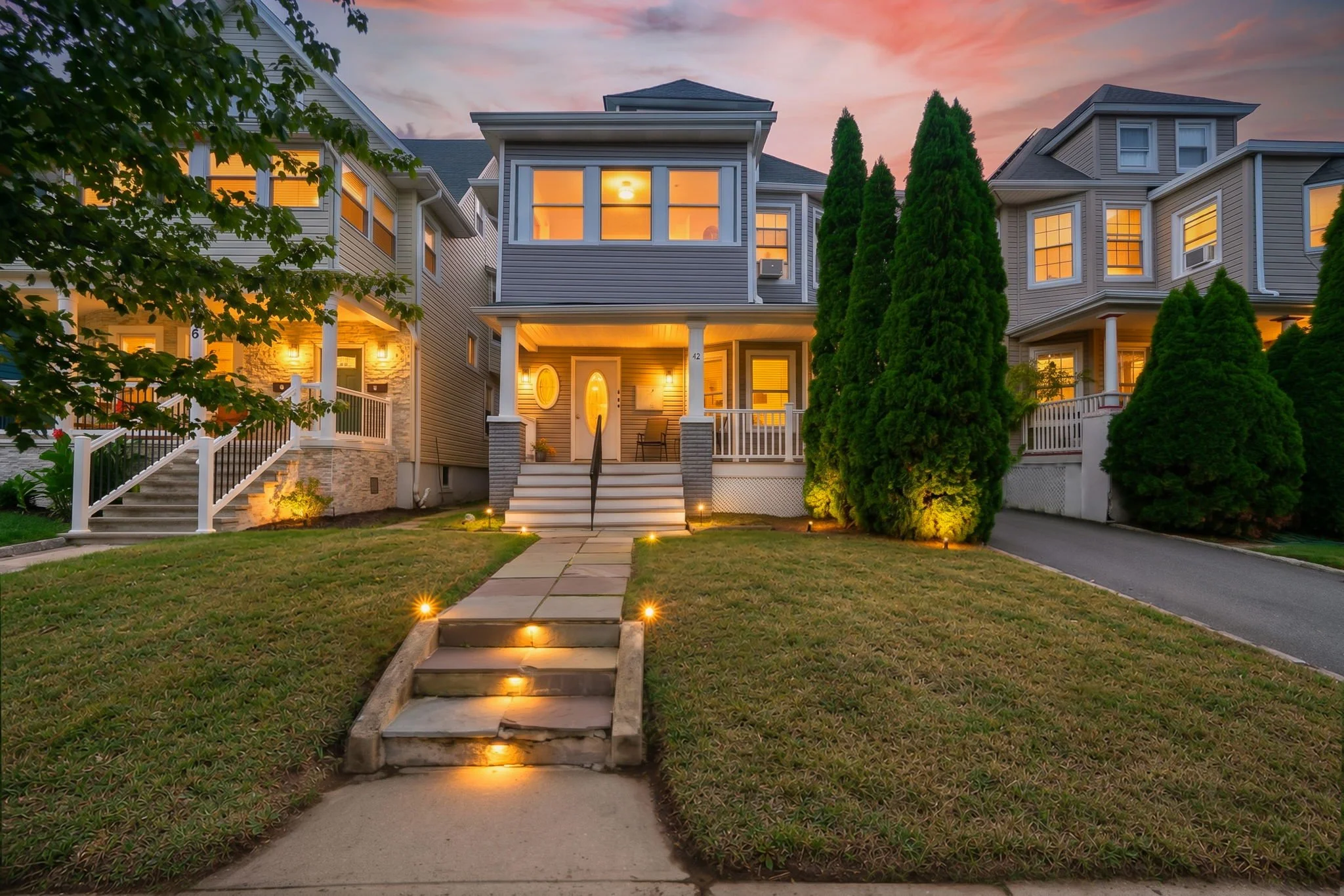 A row of multi-story houses illuminated at dusk with bright yellow lighting visible through windows, a well-manicured lawn, a pathway lit by small outdoor lights, and tall evergreen bushes in the front yard.