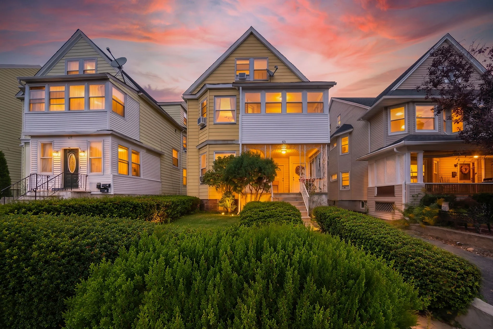 A row of three colorful Victorian-style houses during sunset with warm yellow interior lights, surrounded by green bushes and trees.