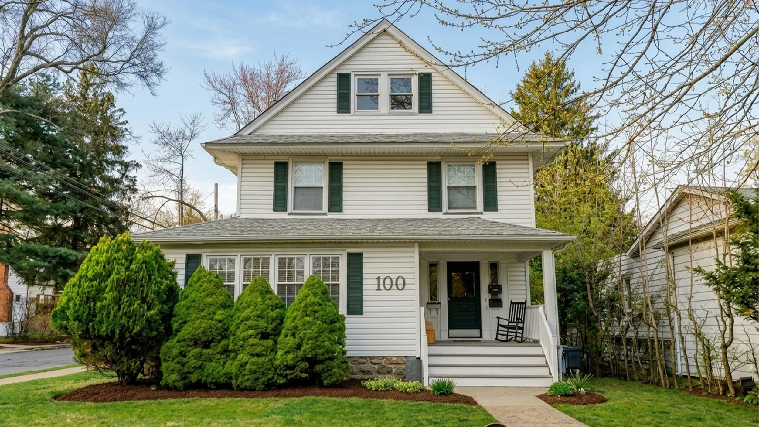 A white three-story house with green shutters, a small front porch, and the number 100 next to the front door. The house is surrounded by green bushes and trees, with a clear blue sky in the background.