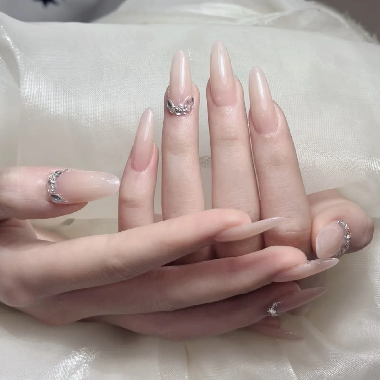 Close-up of a woman's hand with long, almond-shaped, nude-colored nails, decorated with rhinestone rings and a small silver ring with a bow design.