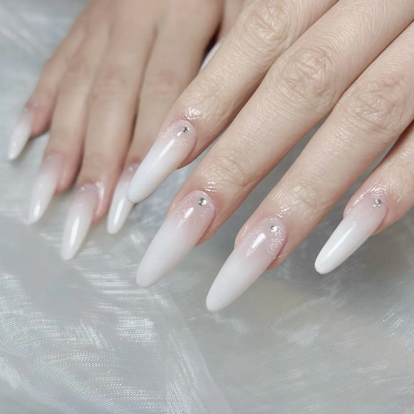 Close-up of hands with long, almond-shaped, white and pink gradient nail extensions decorated with small rhinestones.