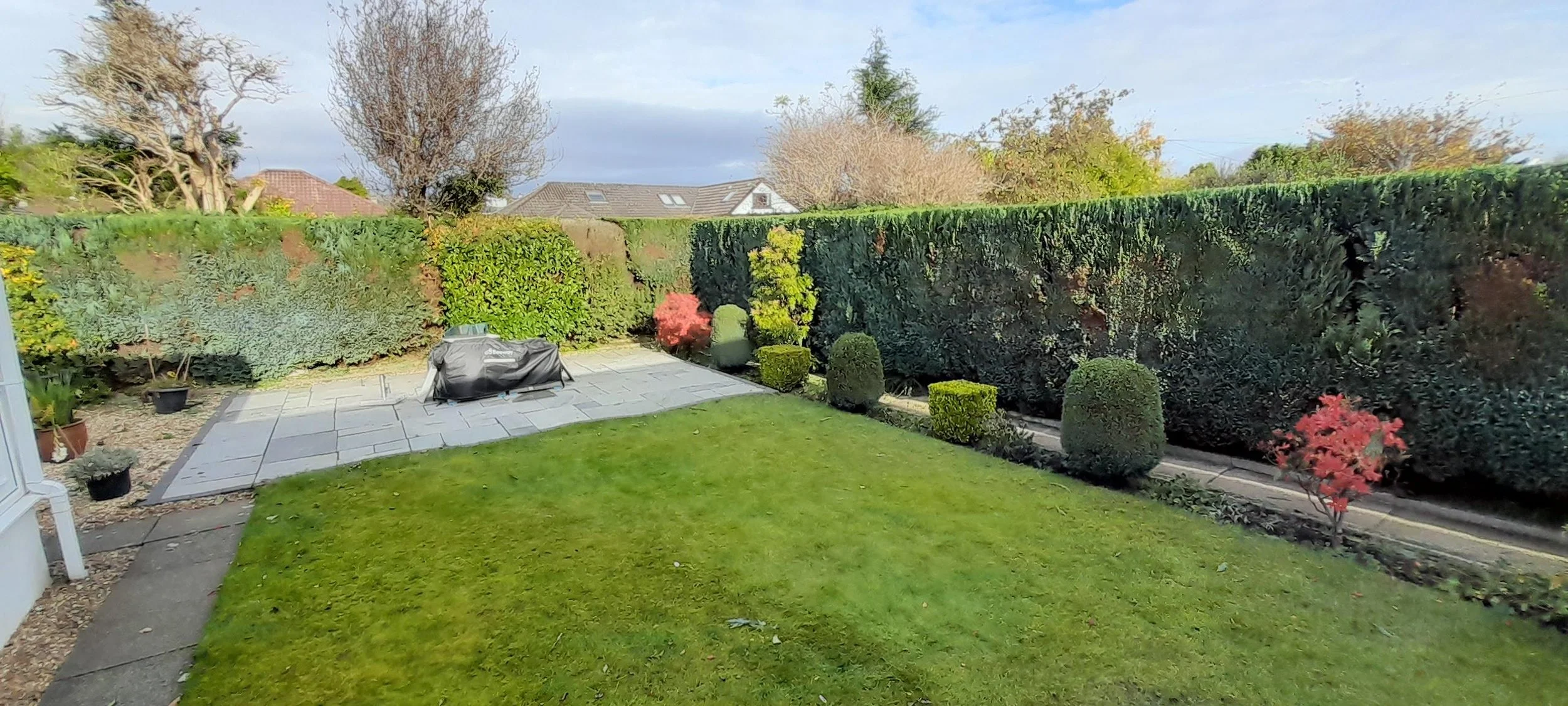 A backyard garden with trimmed hedges, various plants, and a small patio area with a covered grill. The lawn is green and well-maintained, with some landscaping on the edges. In the background, there are trees and rooftops of nearby houses under a partly cloudy sky.