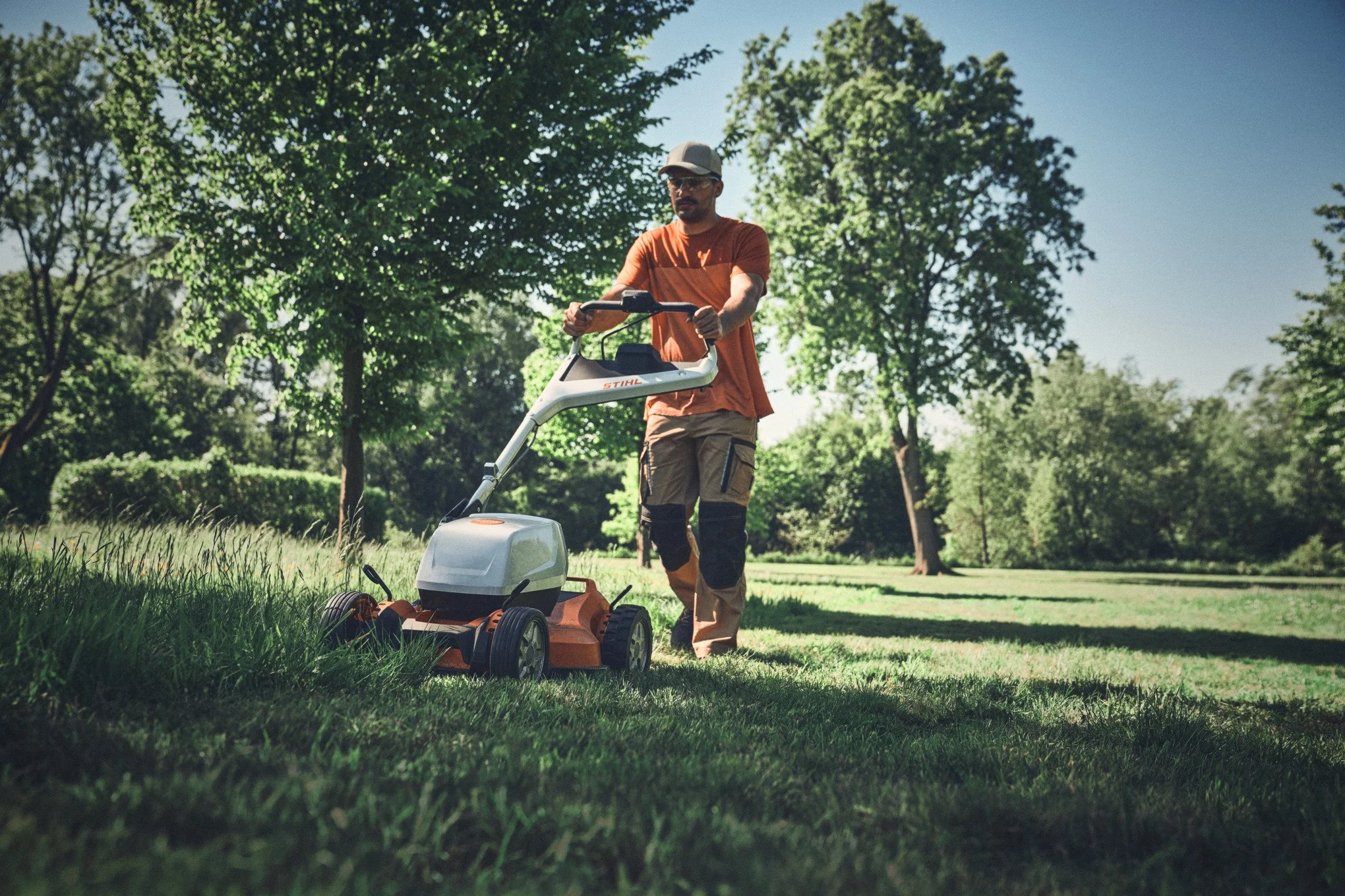 A man mowing a lawn with a power lawn mower in a park-like setting with grass, trees, and a clear blue sky.