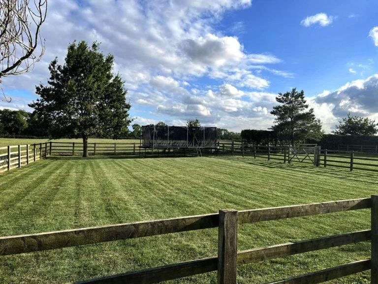 A grassy backyard with a fenced perimeter, a large tree, and a trampoline in the distance under a partly cloudy sky.