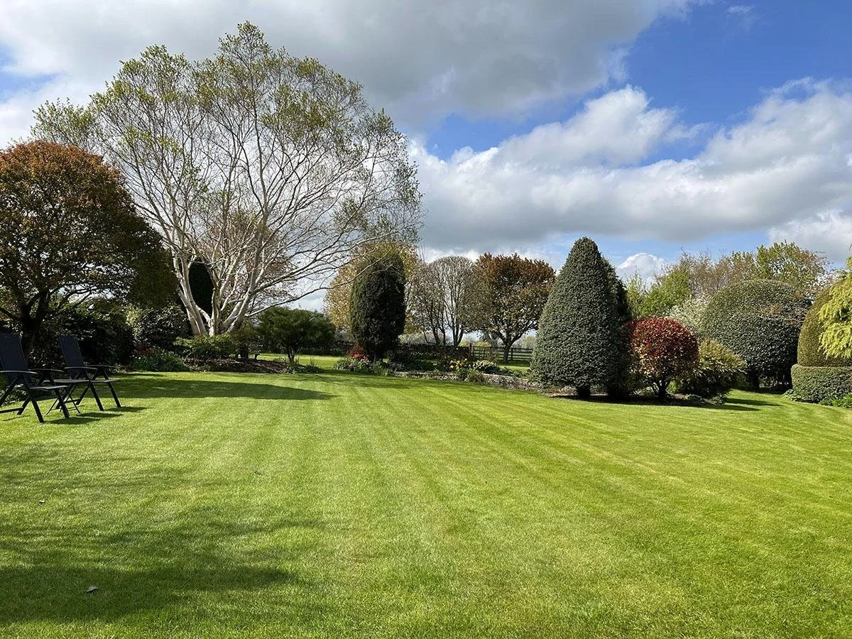 Beautiful landscaped garden with lush green grass, various trees, and shrubs under a partly cloudy sky.
