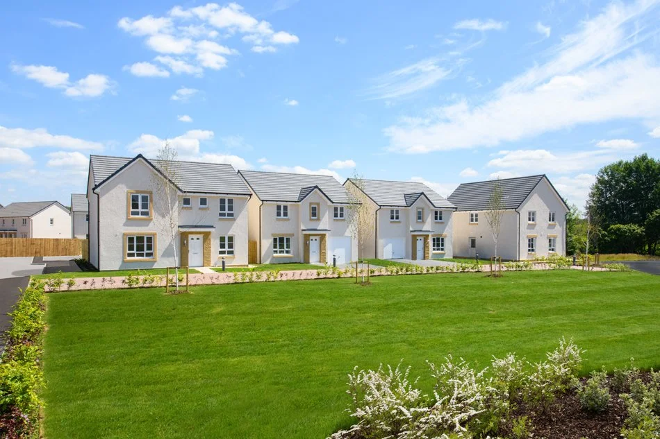 A row of modern townhouses with white exteriors and gray roofs, set in a landscaped area with green grass, small trees, and flower beds under a bright blue sky with clouds.