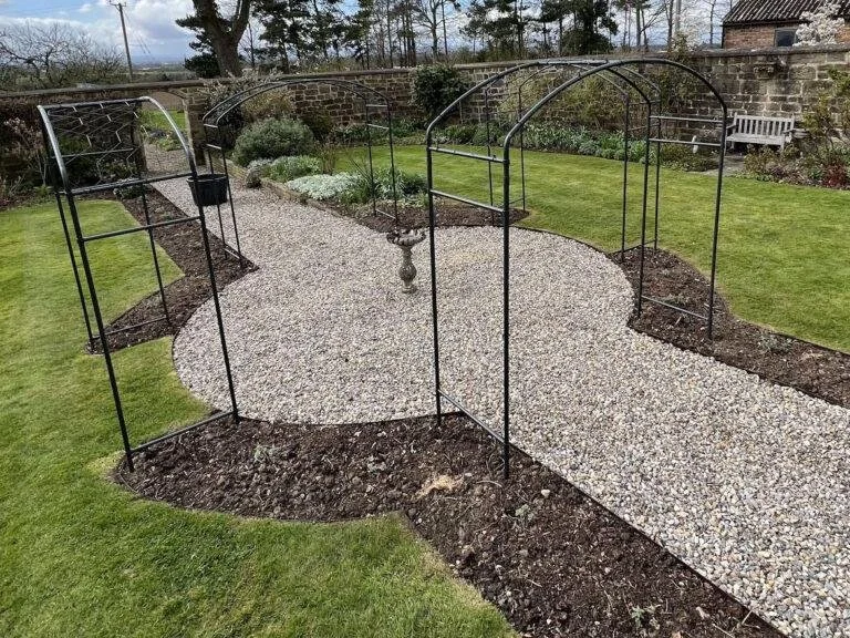 A backyard garden with a gravel pathway, metal arch supports, and some small plants and bushes along the sides.